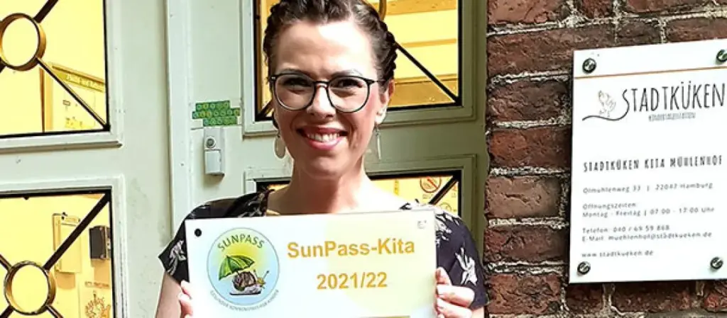 Frau mit Auszeichnung „SunPass-Kita 2021/22“ vor dem Eingang der Stadtküken Kita