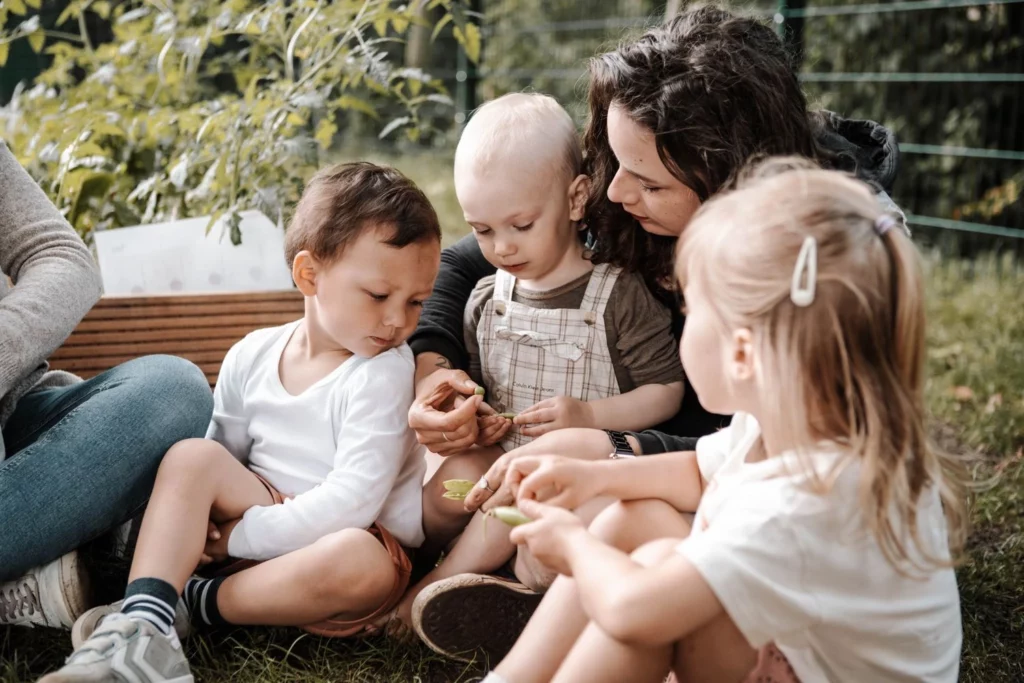 Erzieherin sitzt mit Kindern im Garten und zeigt ihnen Pflanzen zum Entdecken