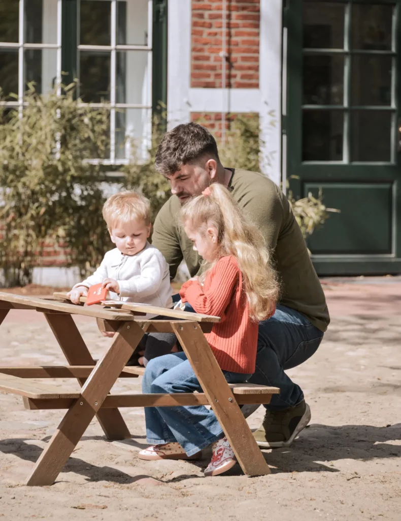 Vater begleitet zwei Kinder beim gemeinsamen Spielen und Entdecken an einem Picknicktisch im Außengelände der Kita.