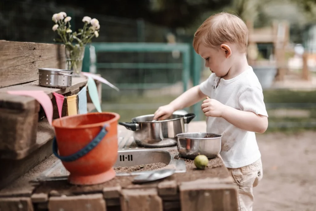 Toddler playing at an outdoor mud kitchen, stirring soil and natural materials in metal pots during imaginative play.