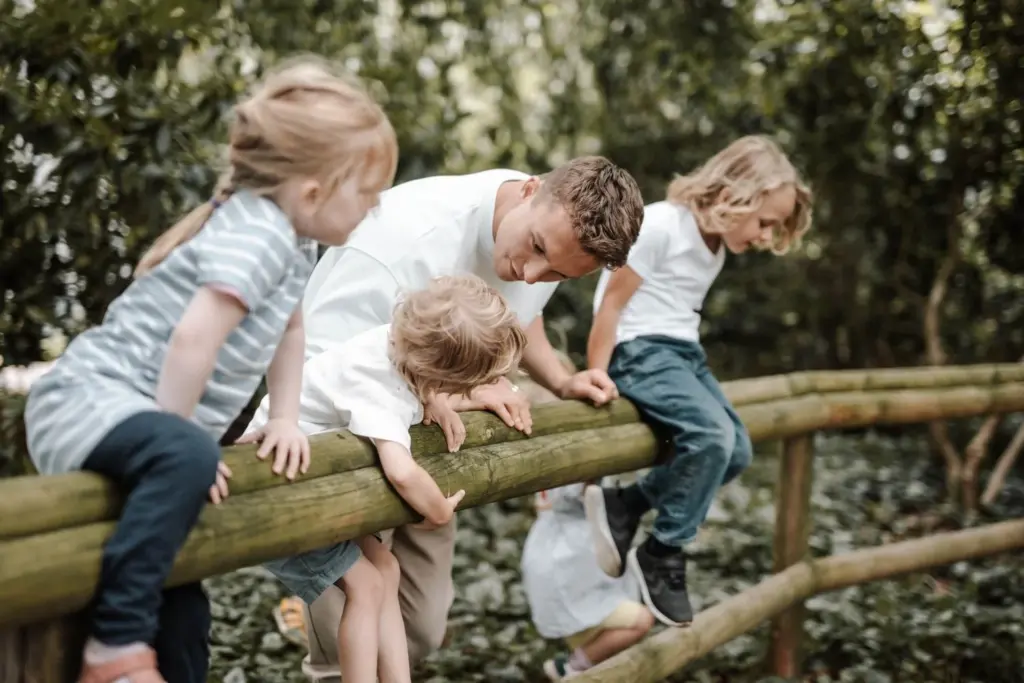 An adult helps several children climb over a log fence in a green space.