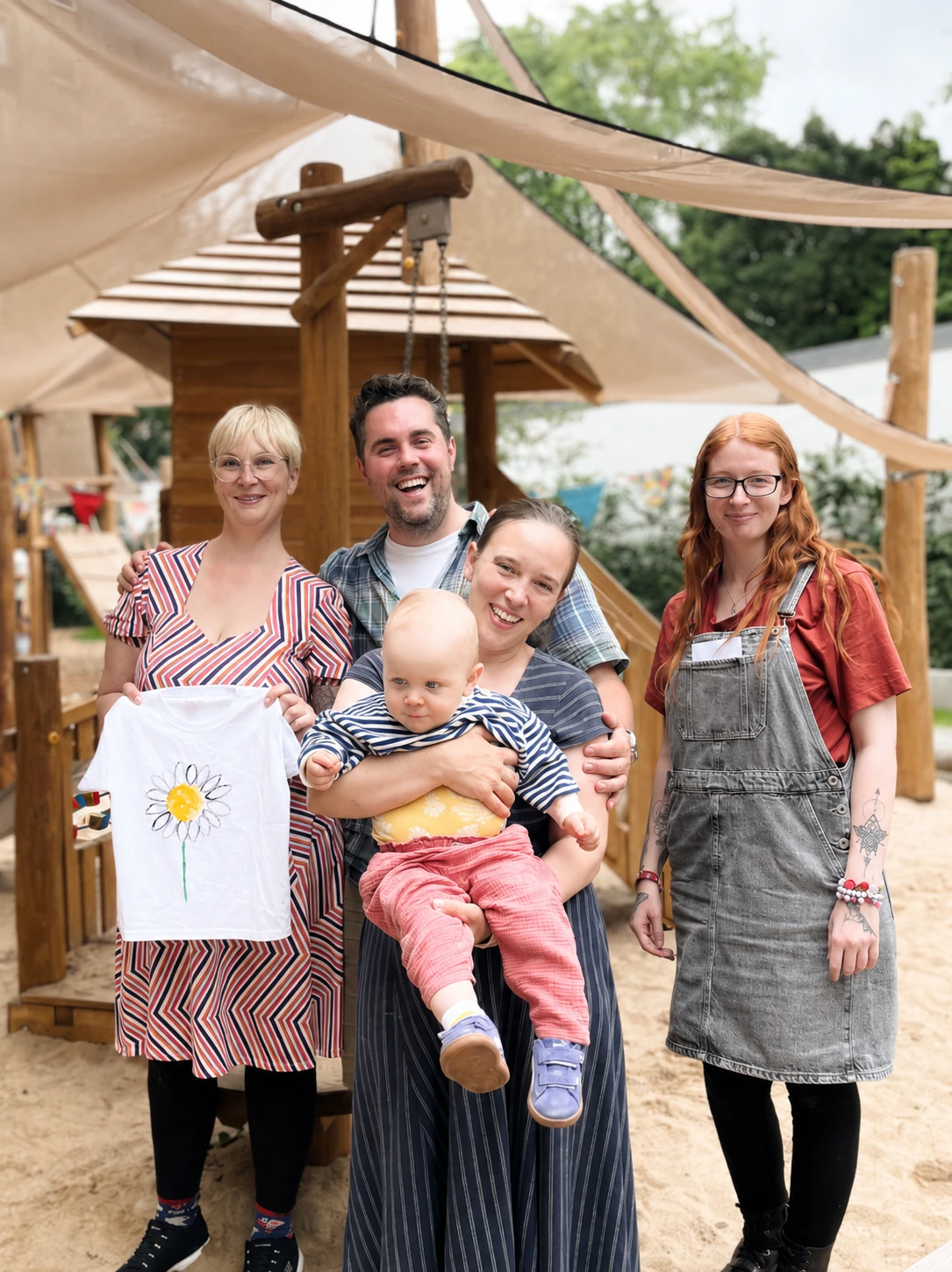 Eine Gruppe von vier Erwachsenen mit einem Baby posiert lachend auf einem Spielplatz.