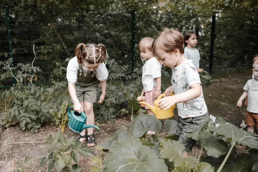 Several children are watering vegetable plants together in a raised bed with colorful watering cans in the kindergarten or school garden.
