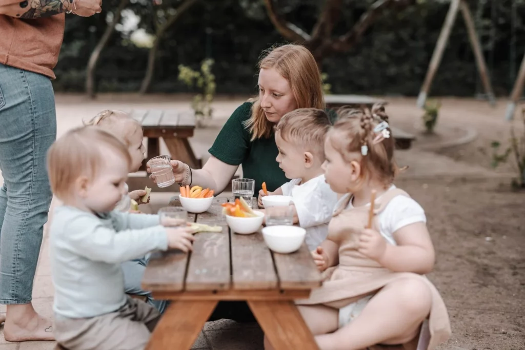 Educator serving water and healthy snacks to toddlers seated together at an outdoor picnic table in the kindergarten garden.